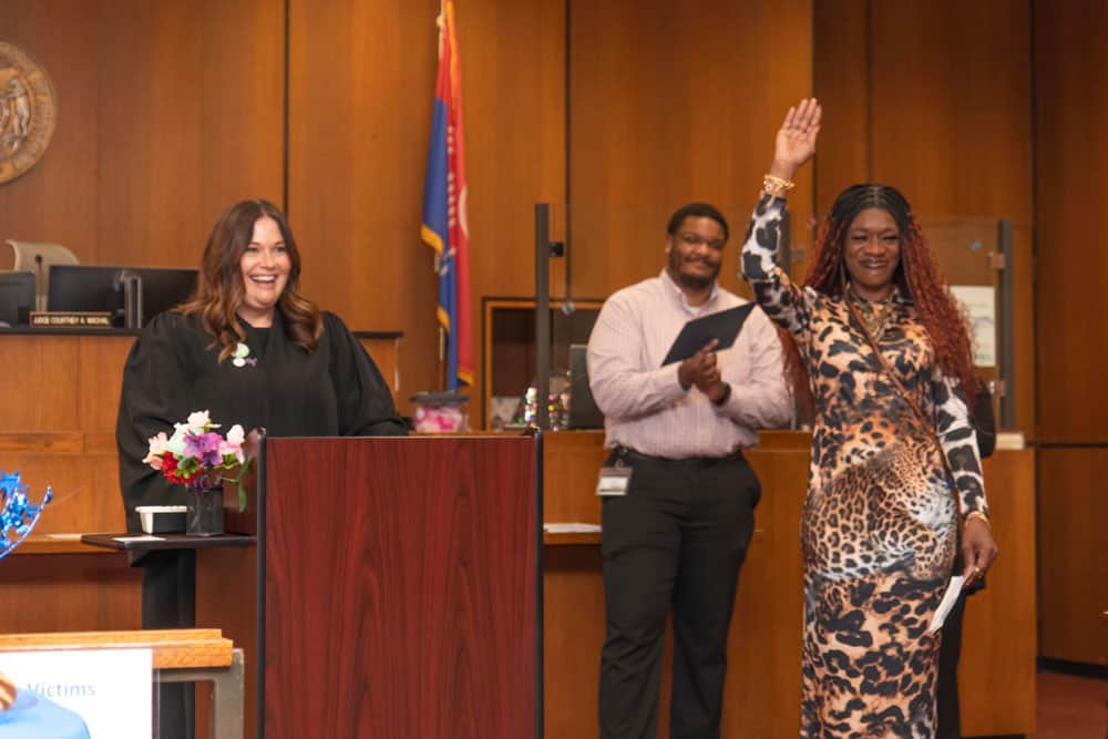 Judge Courtney Wachal smiles from behind a podium while a Wellness Court graduate raises her hand while smiling