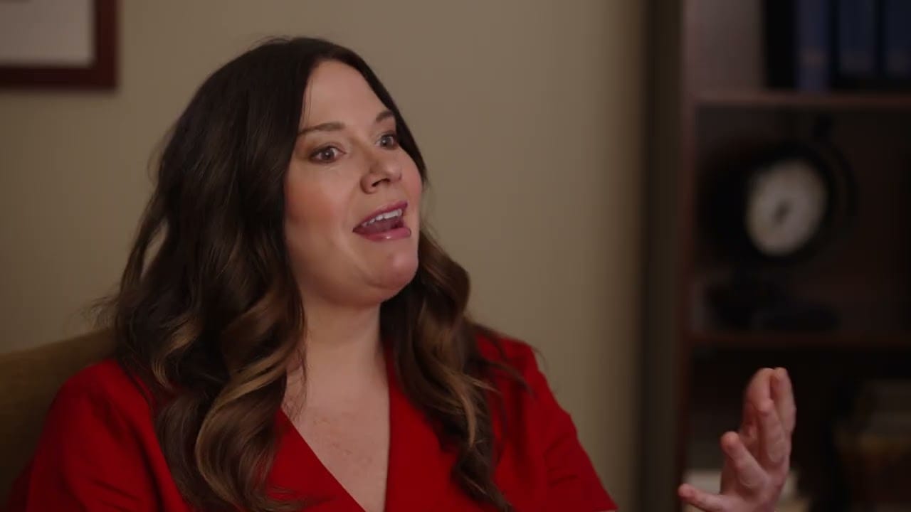 Woman with long brown hair wearing a red top, sitting indoors, gesturing with one hand while speaking. A clock and shelves are visible in the blurred background.