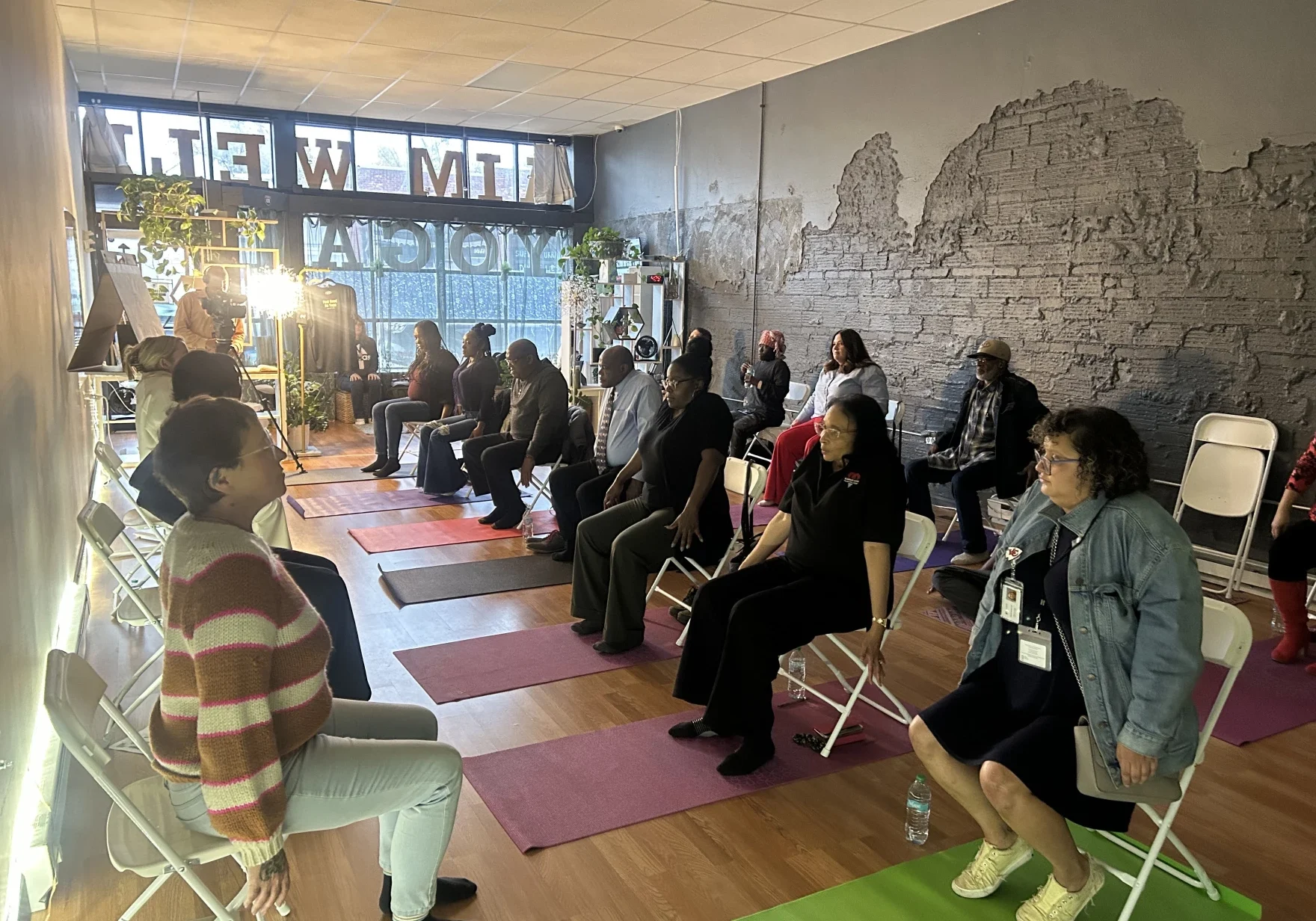 A group of adults sit on chairs arranged in rows inside a room with mats on the floor, participating in a seated exercise or wellness session.